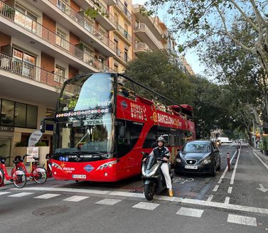 A bright red open-top hop-on hop-off bus in Barcelona, filled with tourists, with landmarks like Sagrada Família and Park Güell in the background under a sunny Mediterranean sky