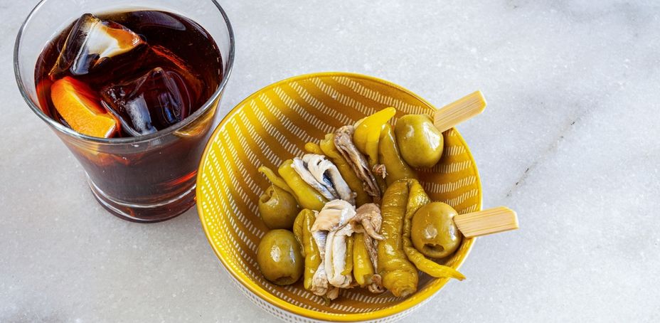 A glass of vermouth with ice and a bowl of Spanish pickled tapas on marble